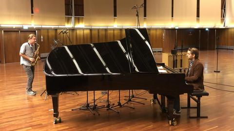 Zwei Musiker in Alltagskleidung spielen in einem Saal mit Parkettboden. Der rechte sitzt am schwarzen, aufgeklappten Konzertflügel, der den größten Teil des Bildes einnimmt. Links davon ein Altsaxofonist.