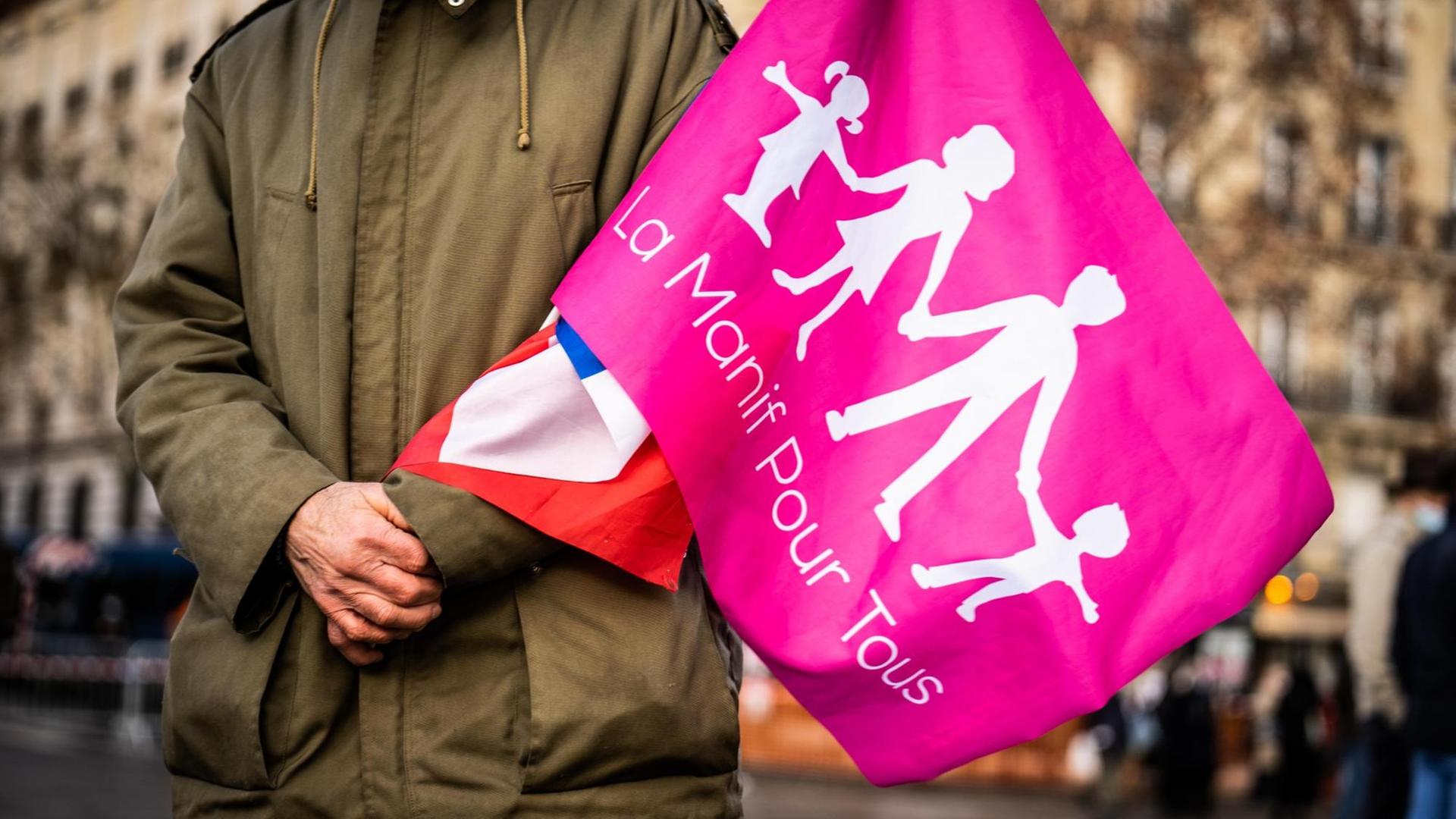 Detail der gekreuzten Hände eines Demonstranten mit einer französischen Flagge und einer Flagge der Demonstration mit einer Darstellung einer Normalfamilie mit Vater, Mutter und zwei Kindern.