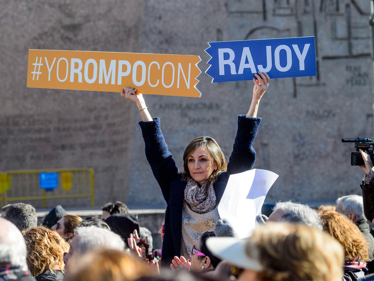 Frau sitzt auf den Schultern von jemandem in einer Menschenmenge und hält Schilder hoch, auf denen auf Spanisch steht: "Ich breche mit Rajoy."