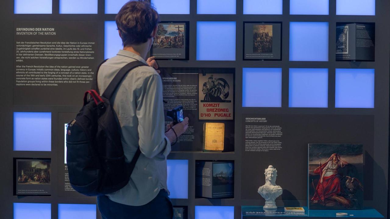 Einblick in die Ausstellung des Dokumentationszentrums Flucht, Vertreibung, Versöhnung im Deutschlandhaus in Berlin. Ein Mann steht vor einer Wand mit kleinen quadratischen drehbaren Schautafel-Würfeln und Schaukästen mit Objekten zum Bereich "Erfindung der Nation" in der Ausstellung.