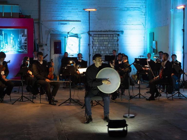 Auf einer Bühne in einem blau ausgeleuchtetem Industrieraum sitzt ein auf Abstand bedachtes Orchester, im Vordergrund ein Mann mit großer runder Trommel.