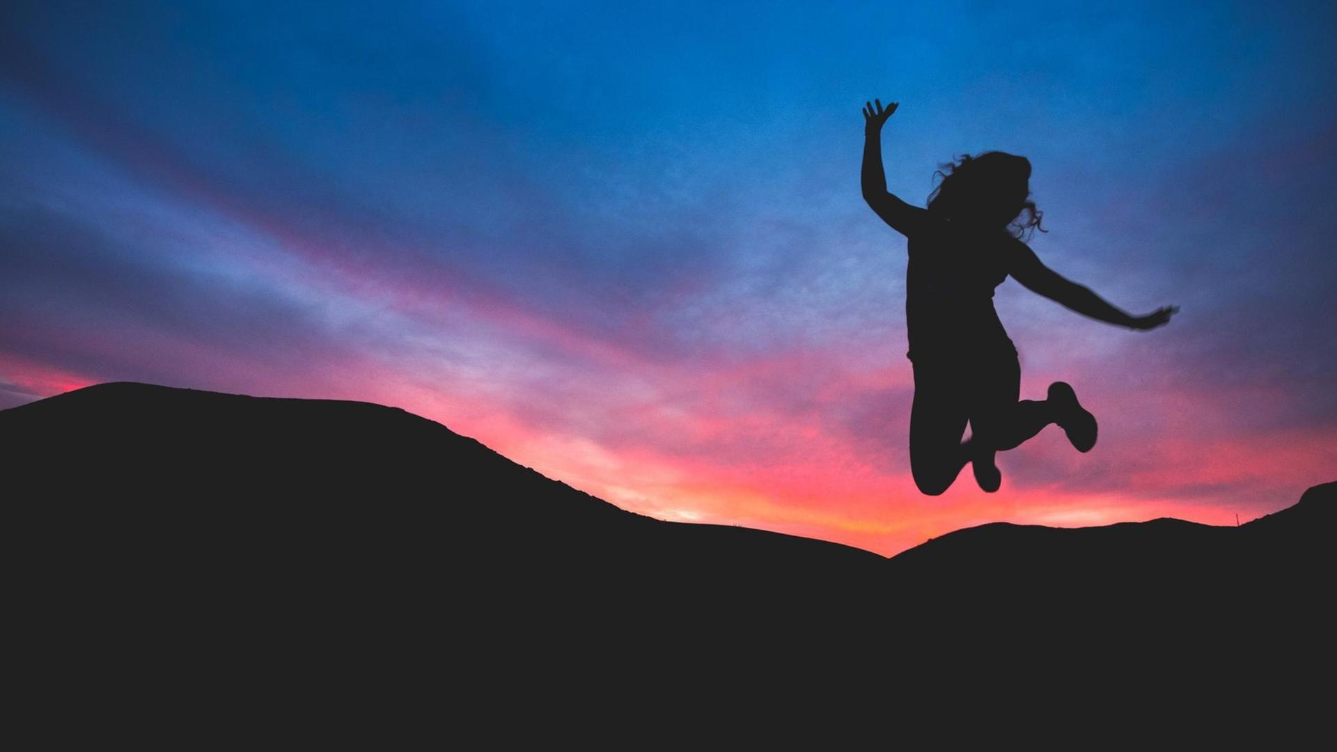 Eine Frau macht einen Luftsprung. Im Hintergrund: Sonnenuntergang und Berge.