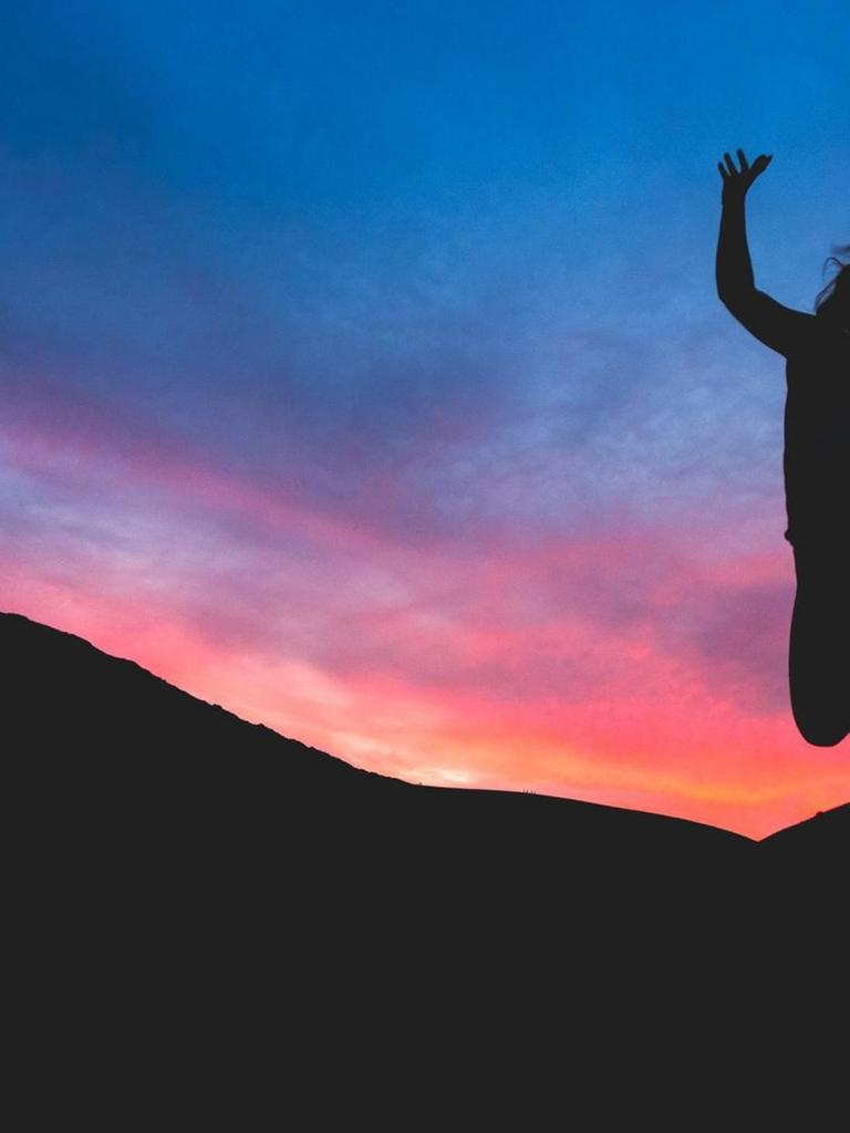 Eine Frau macht einen Luftsprung. Im Hintergrund: Sonnenuntergang und Berge.