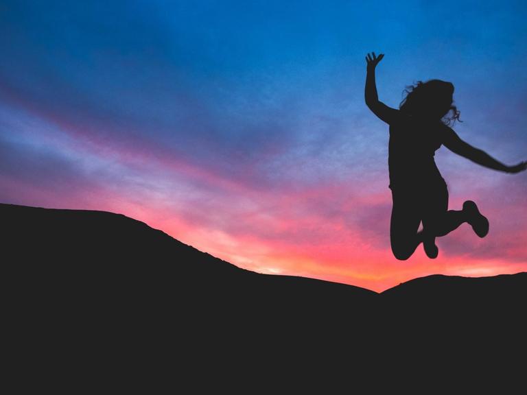 Eine Frau macht einen Luftsprung. Im Hintergrund: Sonnenuntergang und Berge.