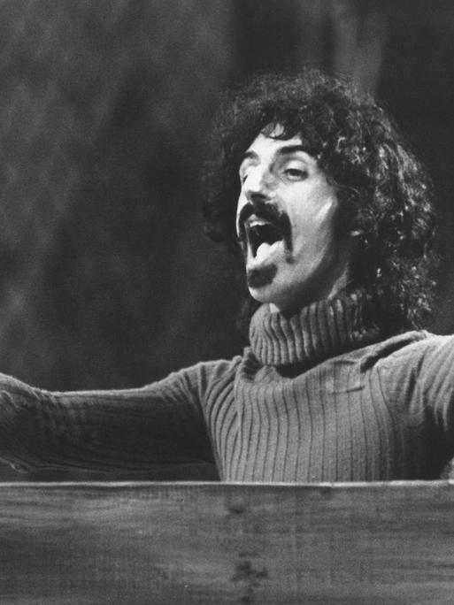 Frank Zappa steht hinter einem Pult in Dirigentenpose.