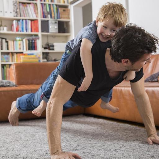 Ein Vater macht Liegestütze im Wohnzimmer mit seinem Sohn auf dem Rücken.