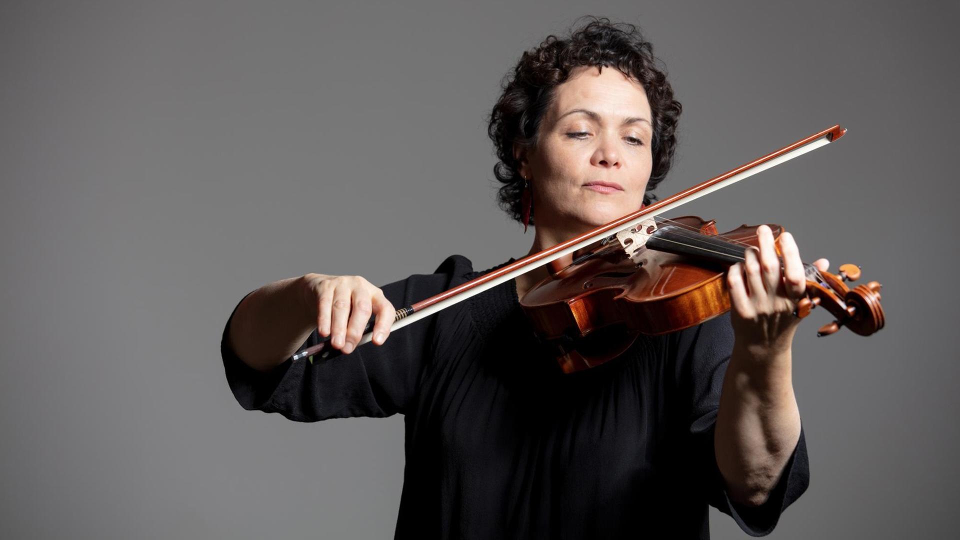 Eine Frau spielt vor einem grauen Hintergrund auf ihrer Geige und schaut auf die Saiten.