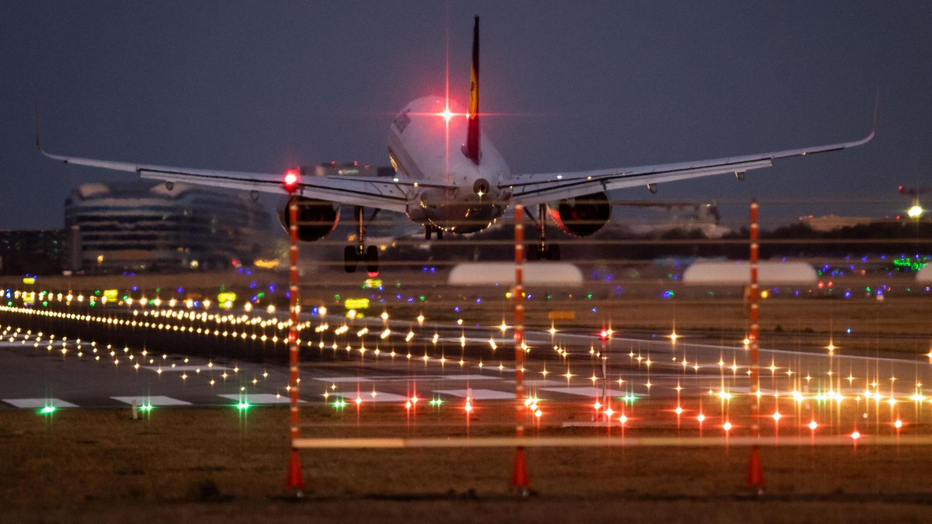 Man sieht das Flugzeug von hinten und im Hintergrund Flughafengebäude. Viele bunte Lichter leuchten in der Dunkelheit.