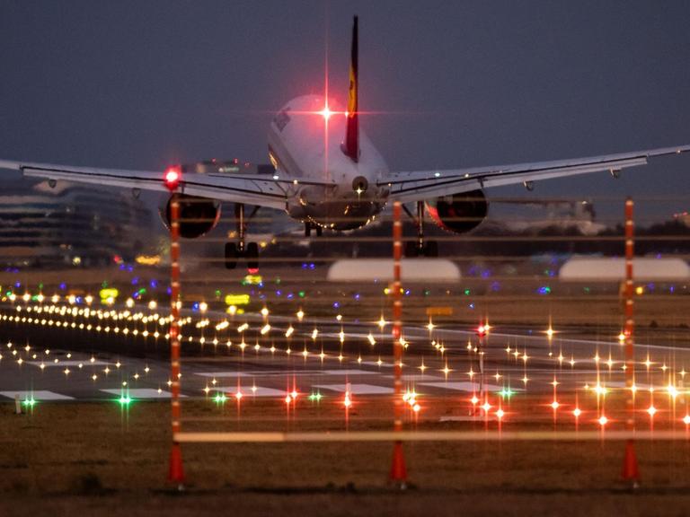Man sieht das Flugzeug von hinten und im Hintergrund Flughafengebäude. Viele bunte Lichter leuchten in der Dunkelheit.