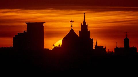 Hinter typisch russischen Kuppeln einer Kirche mit Kreuzen, die sich als schwarze Silhouette ezigen, geht dirket die Sonne glutrot unter.