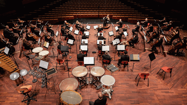 Auf einer Bühne, deren Holz rötlich schimmert, ist ein Orchester mit großen Abständen platziert und spielt vor leerem Auditorium