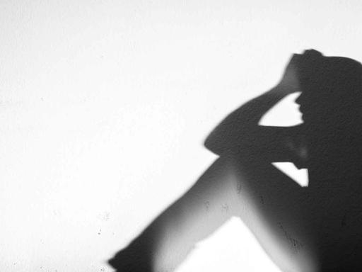 Ein Mensch sitzt auf einer Fensterbank und stützt seinen Kopf mit den Armen, im Gegenlicht sind nur seine Umrisse erkennbar.