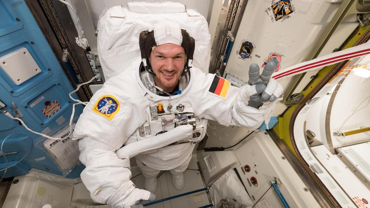 Alexander Gerst hat einen Astronautenanzug an und zeigt mit der linken Hand das "Victory"- bzw. "Peace"-Zeichen.