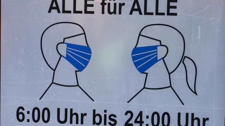 Ein Schild mit der Aufschrift "Alle für Alle - 6:00 bis 24:00 Uhr" weist auf die Maskenpflicht hin.
