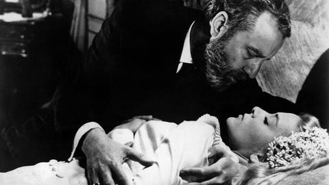 Ein schwarz-weißes Film-Still zeigt einen Mann mit dunklen Haaren und Vollbart, im rechten Arm eine auf einem Bett liegende blonde, jüngere Frau mit geschlossenen Augen haltend