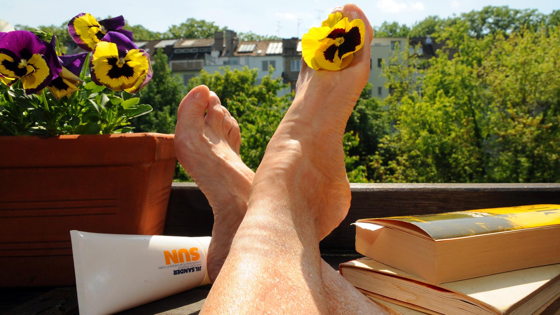 Eine Frau sitzt mit Büchern und Sonnencreme auf einem Balkon und streckt ihre Beine der Sonne entgegen.