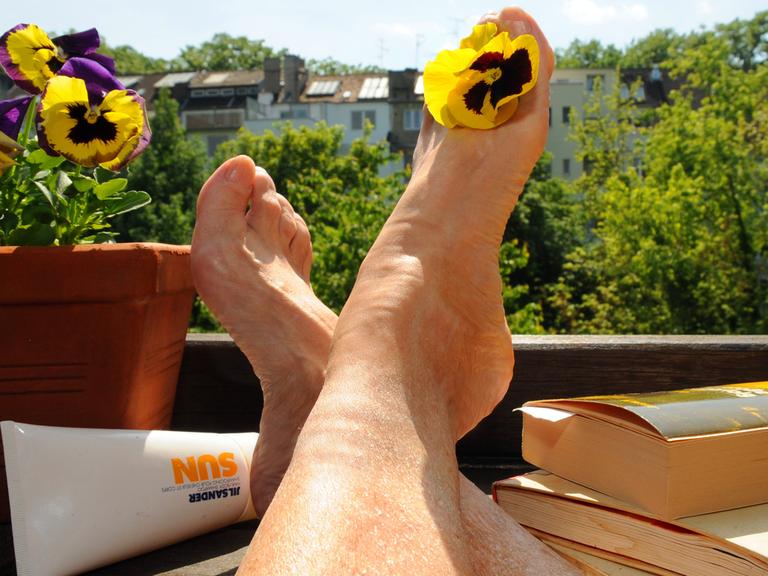 Eine Frau sitzt mit Büchern und Sonnencreme auf einem Balkon und streckt ihre Beine der Sonne entgegen.