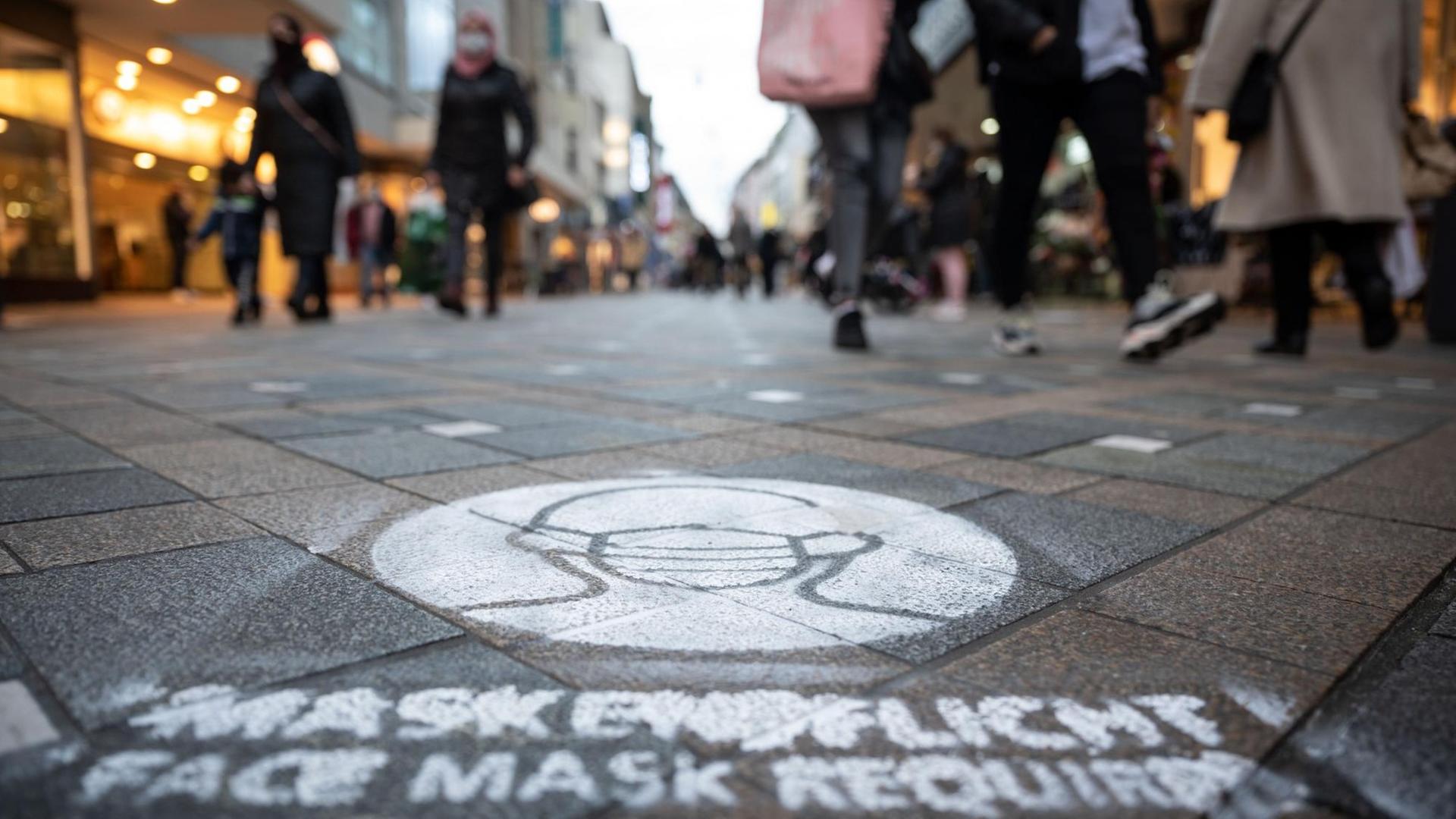 Ein auf den Boden gespraytes Symbol weist in der Fußgängerzone auf die Pflicht hin, Mund-Nasenschutz zu tragen.