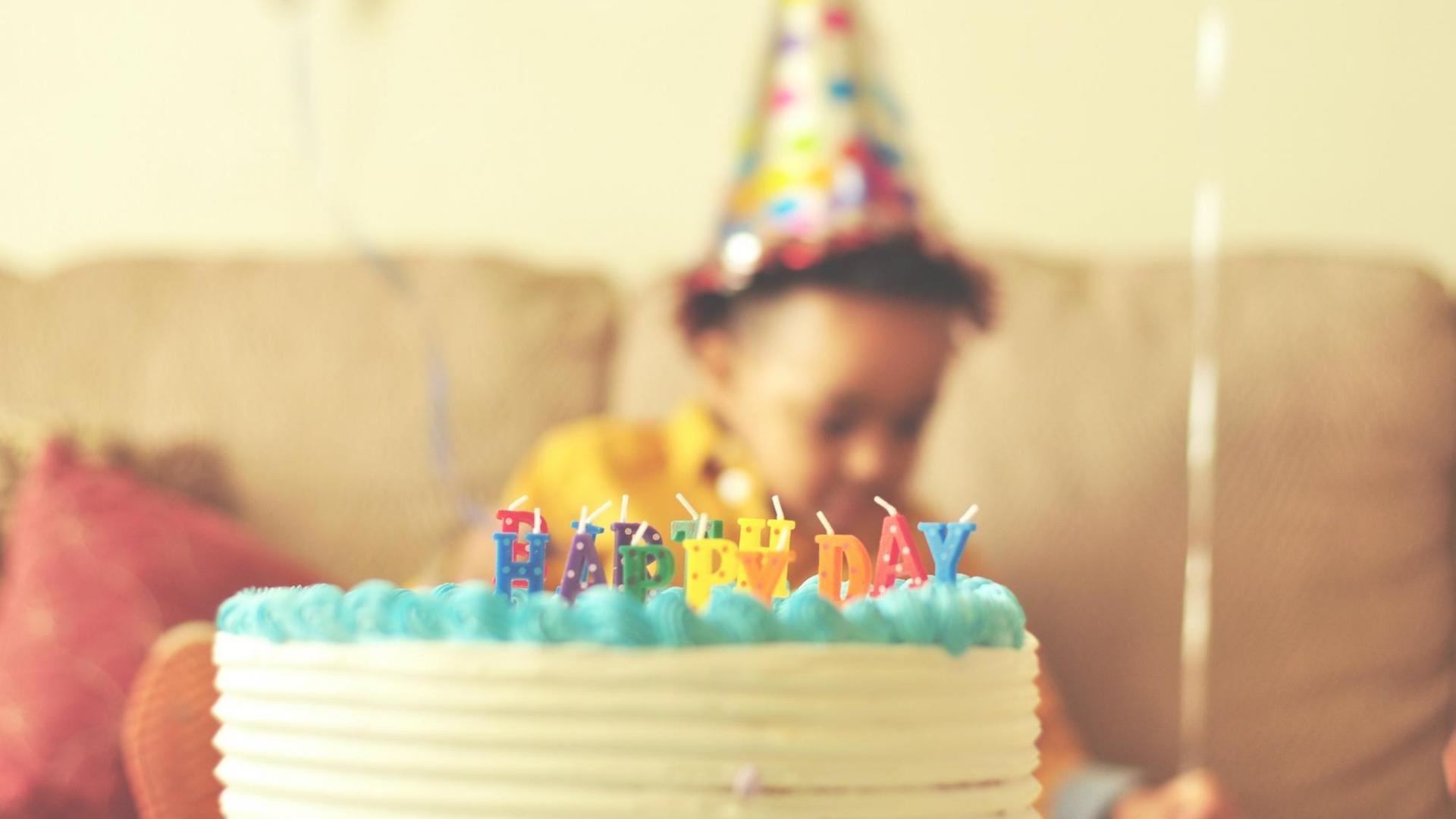 Eine Geburtstagstorte mit "Happy Birthday" in Kerzenbuchstaben. Im Hintergrund ein Kind mit Partyhut.