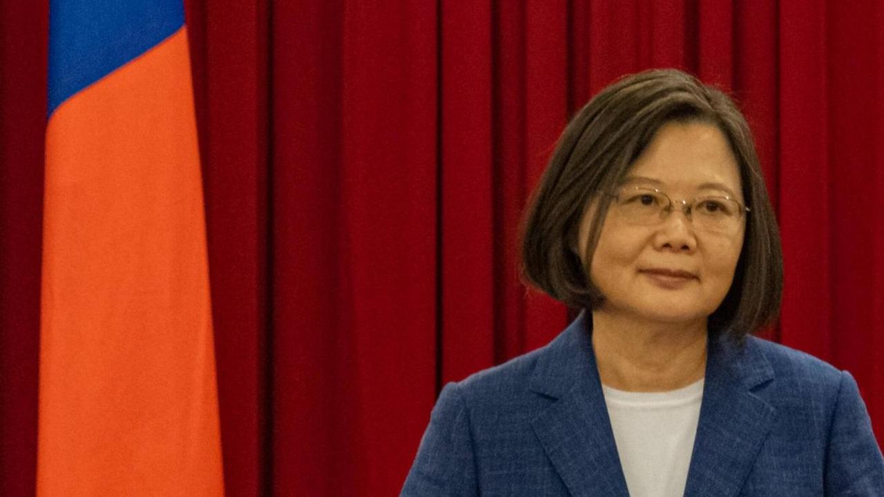 Taiwans Präsidentin Tsai Ing-wen auf einem Podium, neben ihr ist die Flagge Taiwans zu sehen. Sie trägt einen blauen Blazer und ein weißes T-Shirt. Ihre Haare sind dunkel und halblang, sie trägt zudem eine Brille. Im Hintergrund ist ein roter Vorhang zu sehen.