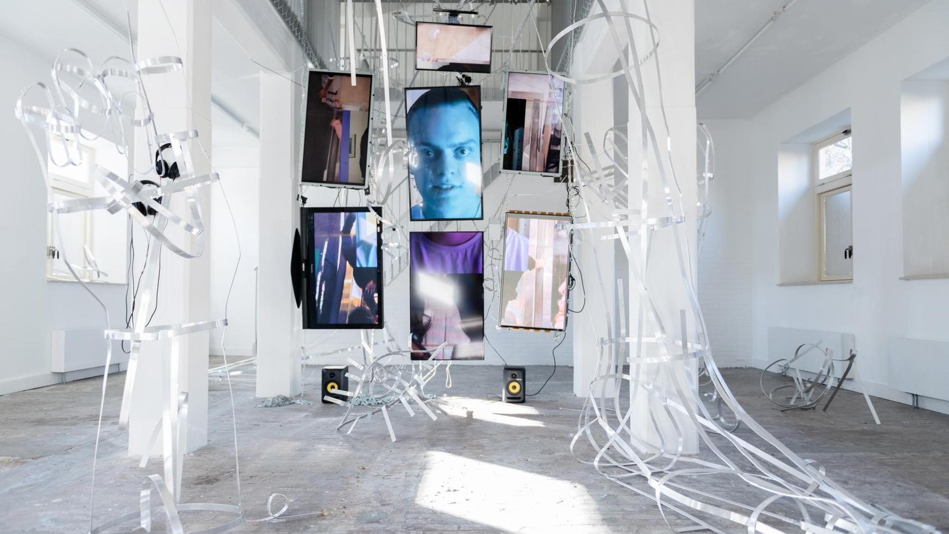 Videoinstallation von Johannes Büttner: Mehrere Bildschirme hängen in einem sehr hohen Raum. Darauf ist ein Männergesicht zu sehen.