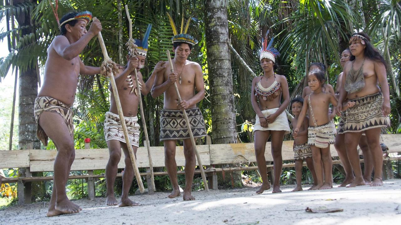 Eine Gruppe von Bora-Indianern steht auf einem sandigen Platz. Die Männer haben lange Holzstäbe in der Hand, während die Frauen und Kinder ihnen zuschauen.
