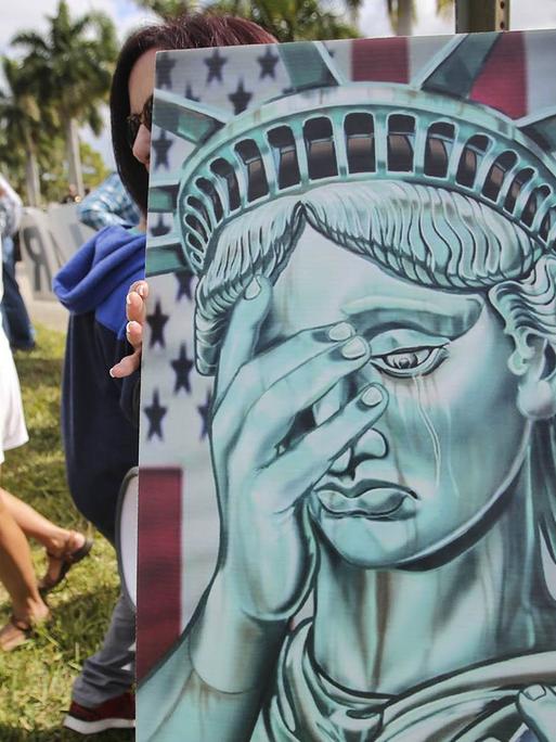 Zu sehen sind Menschen am 21. Januar 2019 vor dem Gemeindehaus in West Palm Beach, Florida. Jemand hält dabei ein Bild mit der Statue of Liberty, die sich eine Hand vor das Gesicht hält.