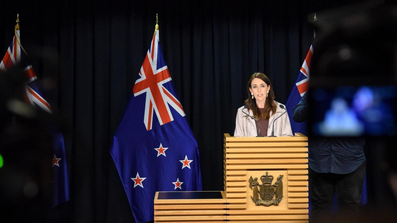 Jacinda Ardern steht an einem Pult und spricht, im Hintergrund ist die Neuseeländische Flagge zu sehen.