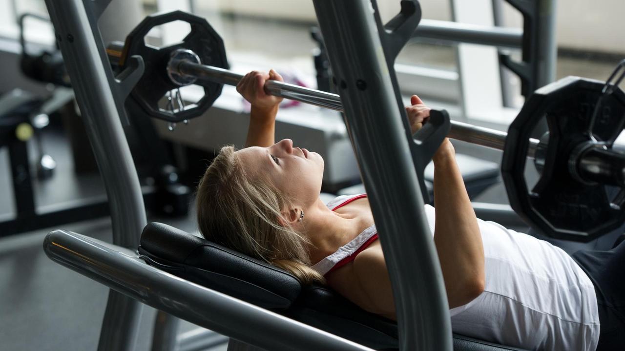 Eine junge Frau in einem Fitnessstudio drückt eine Langhantel auf der Schrägbank in die Höhe. 