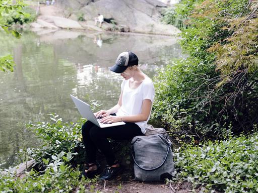 Eine junge Frau sitzt mit ihrem Laptop an einem Seeufer im Grünen