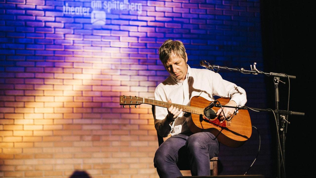 Ein Mann im weißen Hemd sitzt auf einem Hocker und spielt Gitarre. Auf dem Boden steht ein Weinglas.