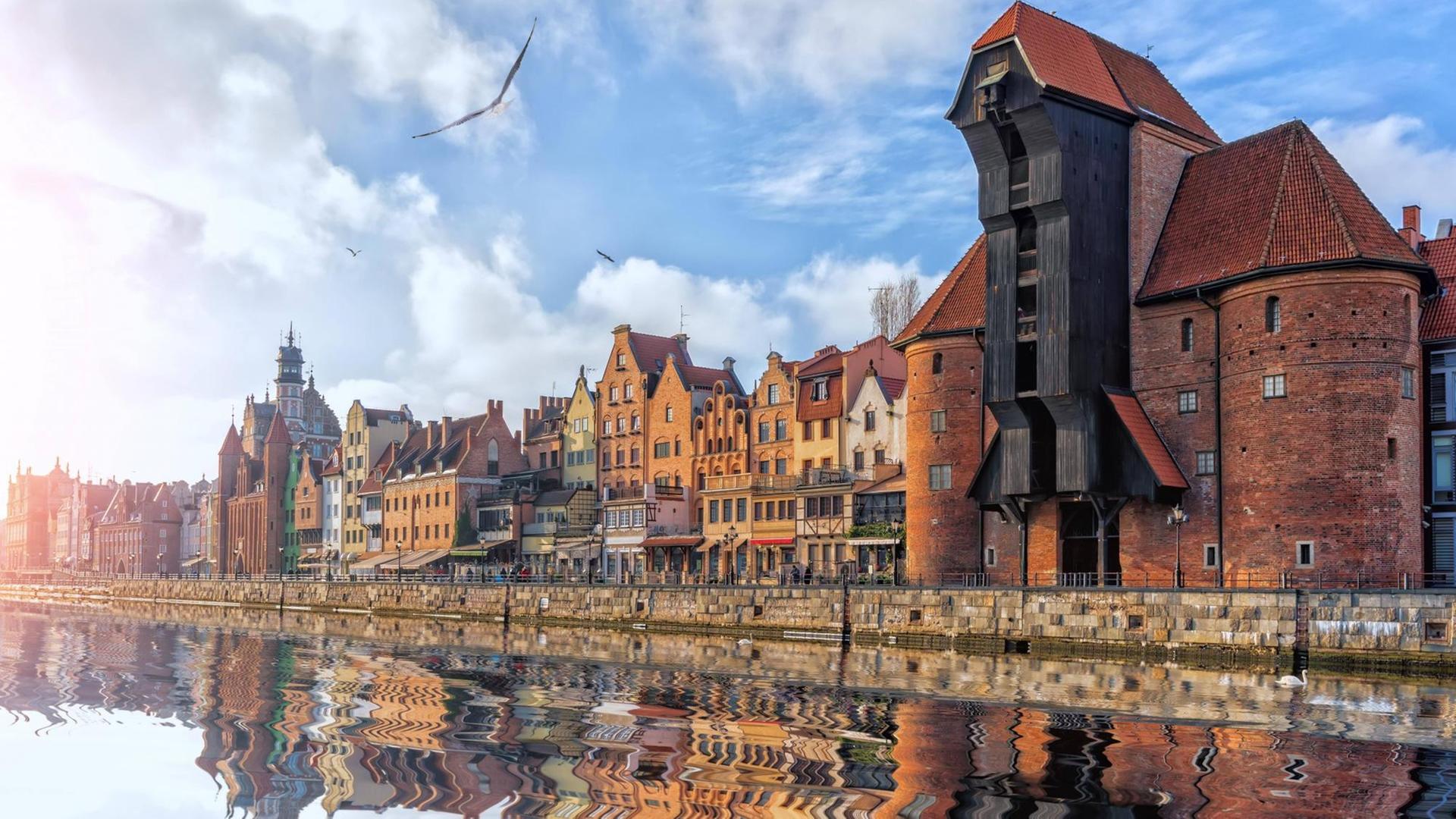 Blick auf das Hafengelände mit historischem Kran und alten Häusern, die sich allesam im Wasser spiegeln.