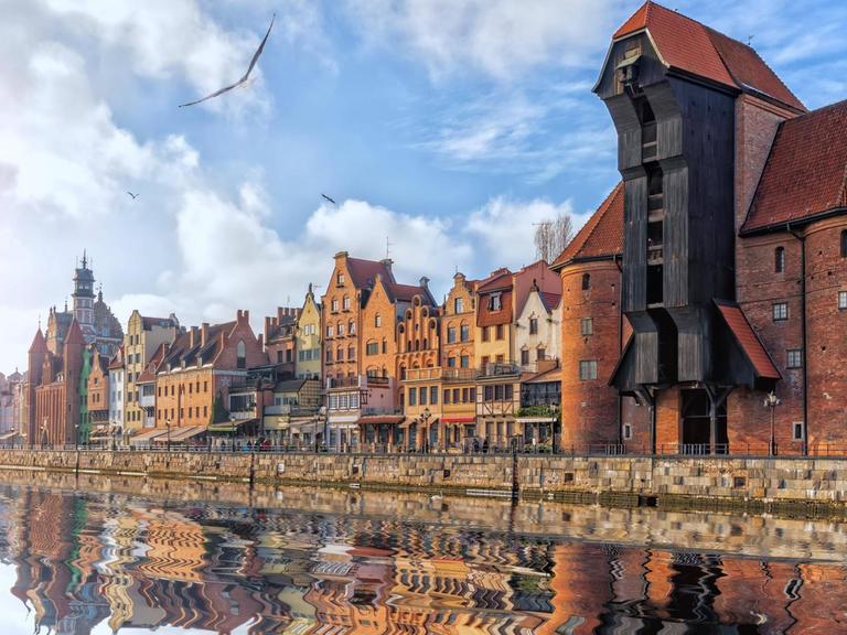 Blick auf das Hafengelände mit historischem Kran und alten Häusern, die sich allesam im Wasser spiegeln.