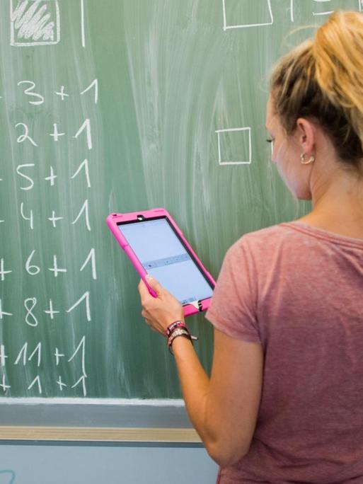 Eine Lehrerin steht mit einem Tablet in der Hand an der Tafel und schreibt Mathe-Aufgaben an