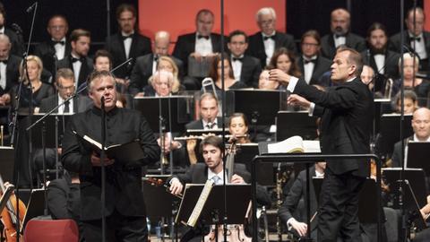 Ein groß besetztes Orchester, dahinter ein Chor. Vorne steht der Dirigent und ein Sänger.