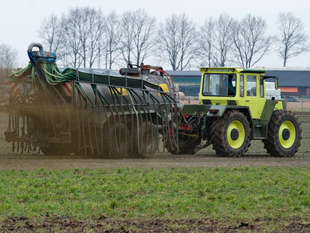 Ein gelber Traktor mit Anhänger, aus dem braue Gülle auf ein Feld gespritzt wird.