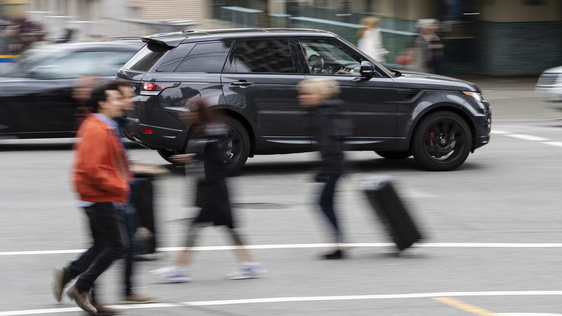 Ein großer, schwarzer Sportgeländewagen fährt in hoher Geschwindigkeit an Fußgängern vorbei durch die Innenstadt.