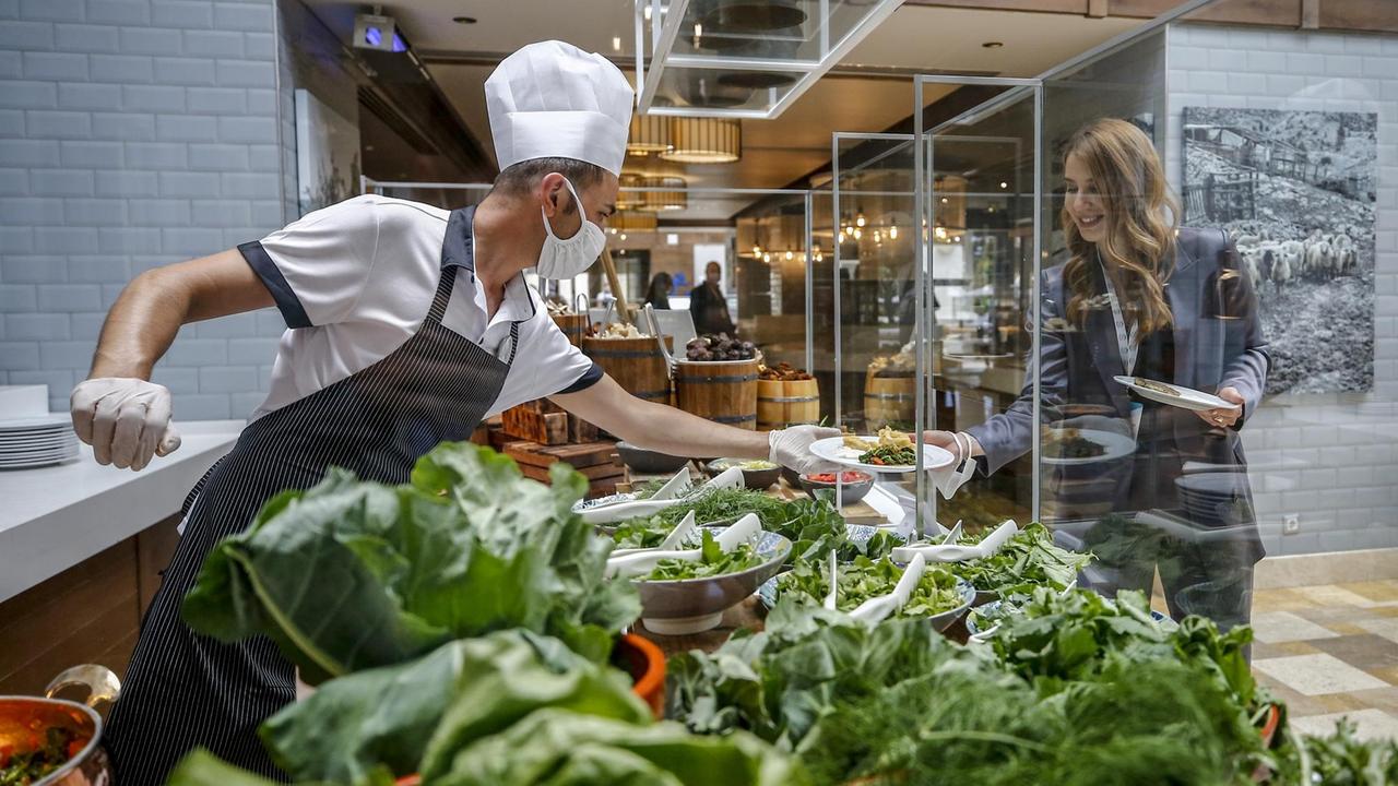 Ein Koch mit Handschuhen und Mundschutz reicht einem Gast in einem Hotel im türkischen Antalya durch eine Durchreiche in einer Scheibe einen Teller mit Essen, aufgenommen im Juni 2020.