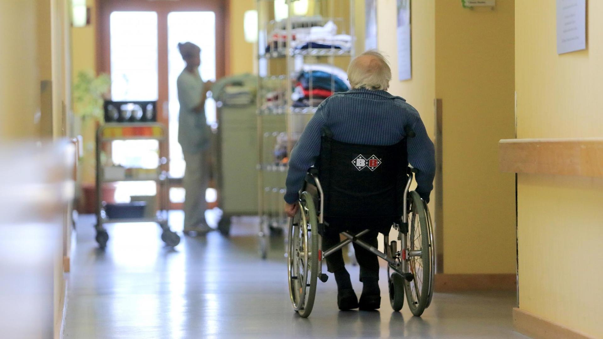 Ein Rentner fährt mit seinem Rollstuhl über einen Flur. Im Hintergrund ist eine Pflegekraft beschäftigt.