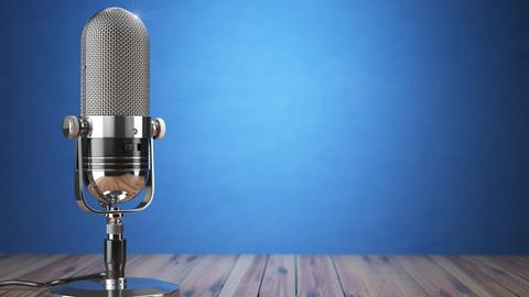 Vor einem blauen Hintergrund steht auf einem Holzboden ein Retro-Mikrofon aus silber-glänzendem Material.