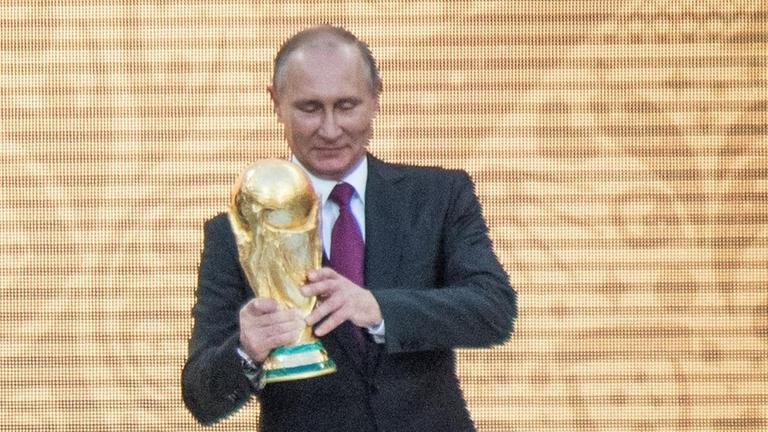 Putin (re.) hält eine goldene Trophäe, Infantino kommt von links auf ihn zu. Vor der Bühne stehen Zuschauer.