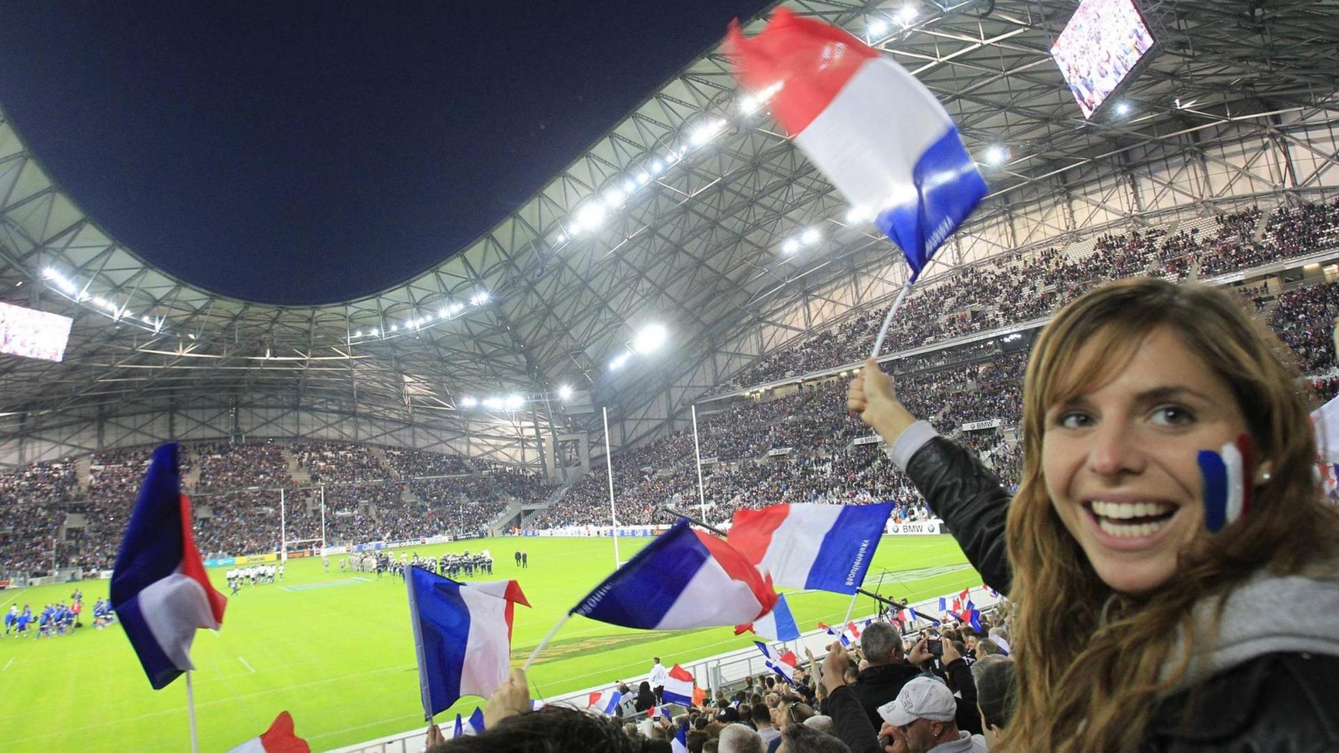 Blick in ein Rugby-Stadion mit Fans, die französische Fähnchen schwenken, im Vordergrund