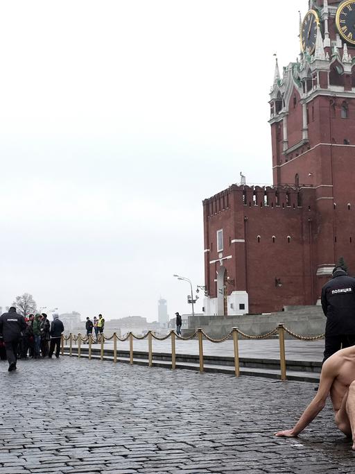 Der russische Aktionskünstler Pjotr Pawlenski sitzt bei der Aktion "Fixierung" nackt auf dem Roten Platz, wo er seinen Hodensack festgenagelt hat. Im Hintergrund die Kremlmauer, die Basilius-Kathedrale, ein Polizist und Touristen.