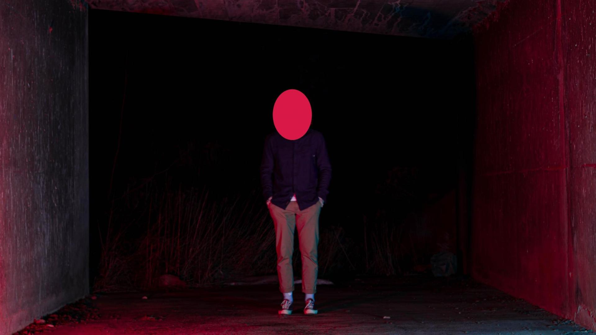 Eine männlich wirkende Person steht zwischen zwei Wänden in rotem Licht. Über dem Kopf ist eine rote elliptische Fläche, so dass kein Gesicht zu erkennen ist.
