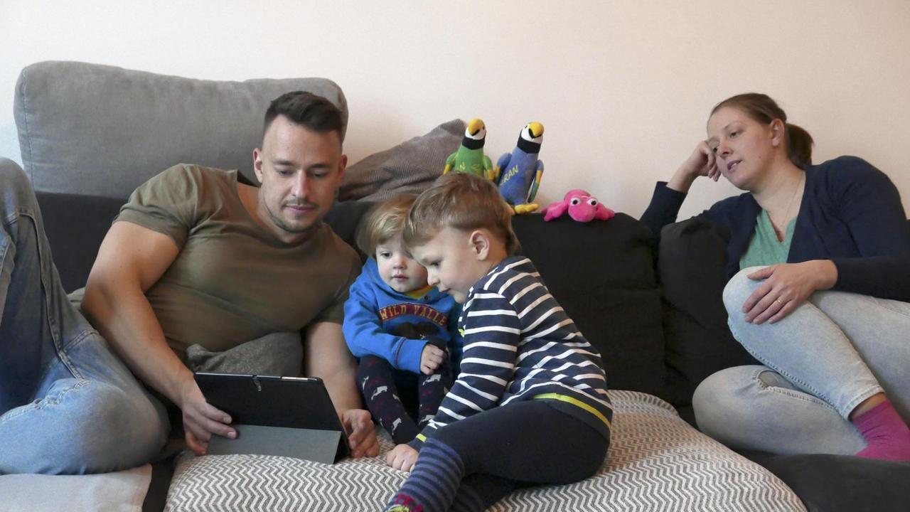 Ein junger Mann und seine zwei Kinder sitzen auf der auf der Couch und schauen auf ein Tablet, eine junge Frau sitzt rechts neben ihnen.