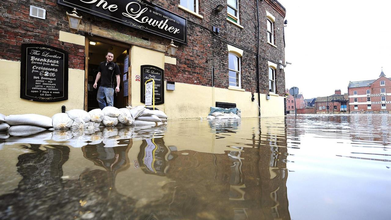 Eine Straße in York, Nordengland, steht durch Überschwemmungen völlig unter Wasser. Auf der linken Seite  steht ein Mann in der Tür seines Pubs.