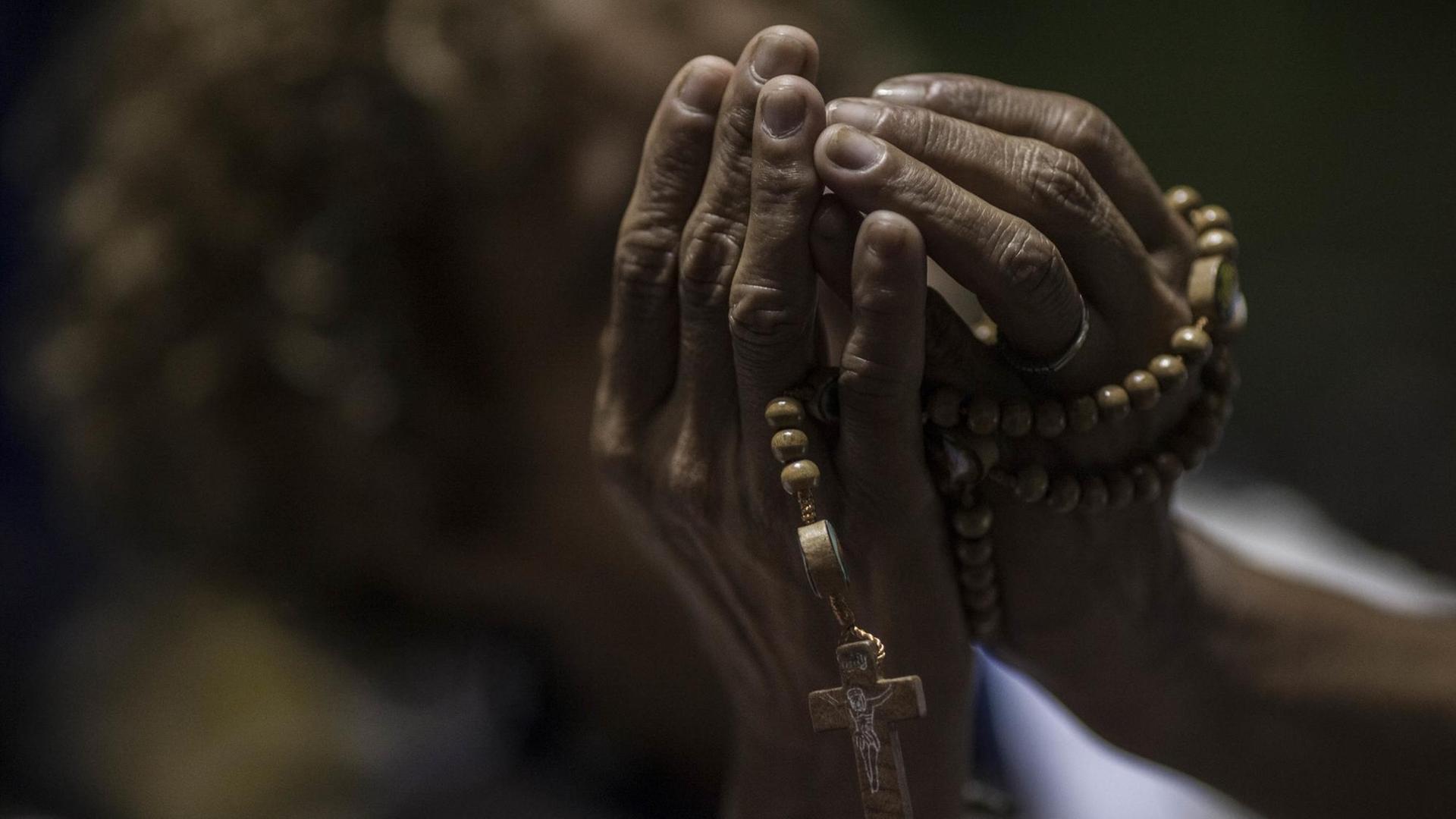 Dunkelhäufige Hände halten beim Gebet eine Kette mit einem Kreuz.