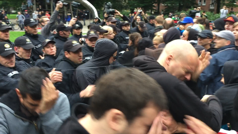In einer Menschenmenge setzt ein Polizist Reizgas ein, mehrere Menschen halten sich die Hände vors Gesicht