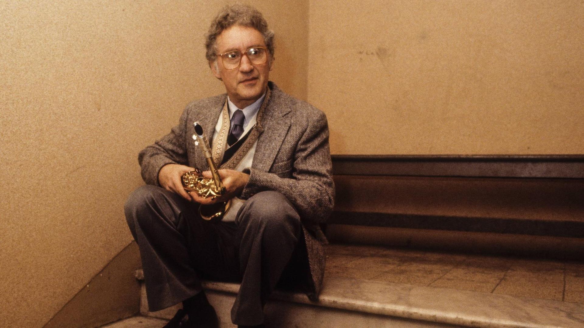 Ein grauhaariger Mann mit Brille sitzt auf einer Stufe in einem Treppenhaus eines Gebäudes. Er trägt einen Anzug und hält sein Altsaxofon auf den Knien.
