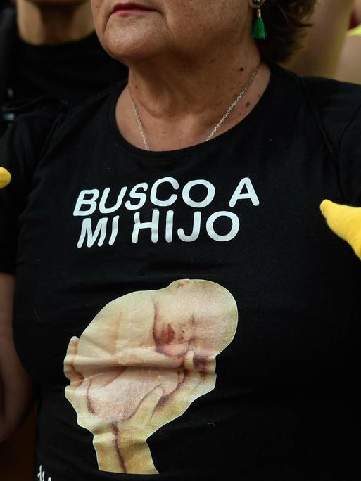 Eine Frau trägt ein schwarzes T-Shirt mit einem aufgedruckten Baby-Foto. Darauf steht: "Ich suche meinen Sohn. Geboren im April 1976 in Cádiz."